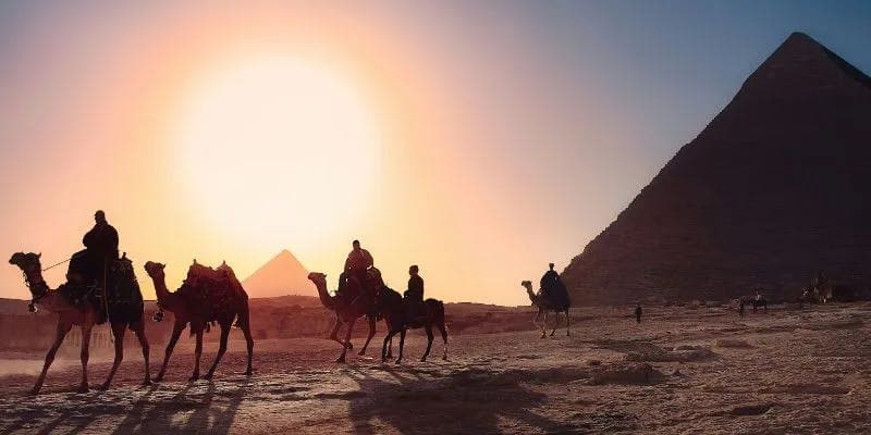 Silhouette of a group of travelers riding camels in a desert near a pyramid, under a large setting sun.