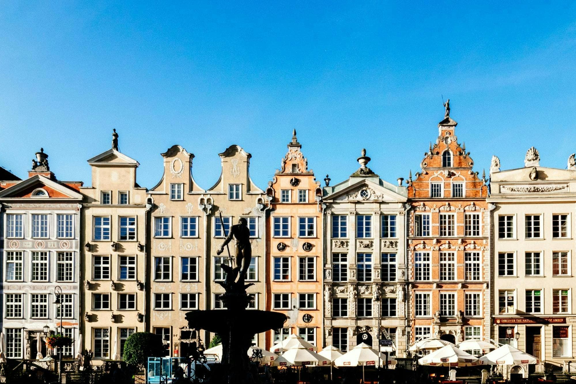 Ornate historic buildings with a central fountain and outdoor café seating under a clear blue sky.