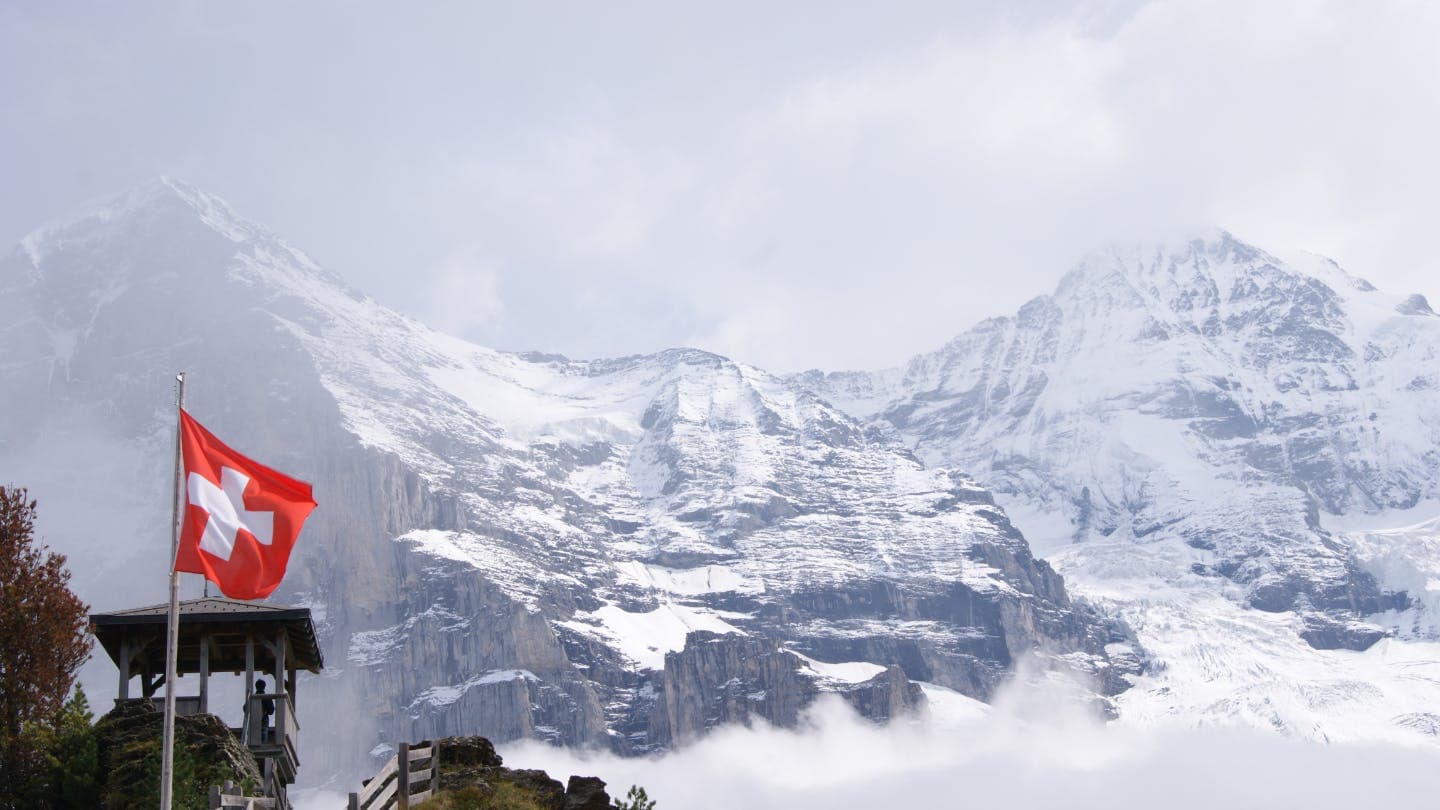 A scenic view of snow-capped mountains in Switzerland with a Swiss flag waving in the foreground.