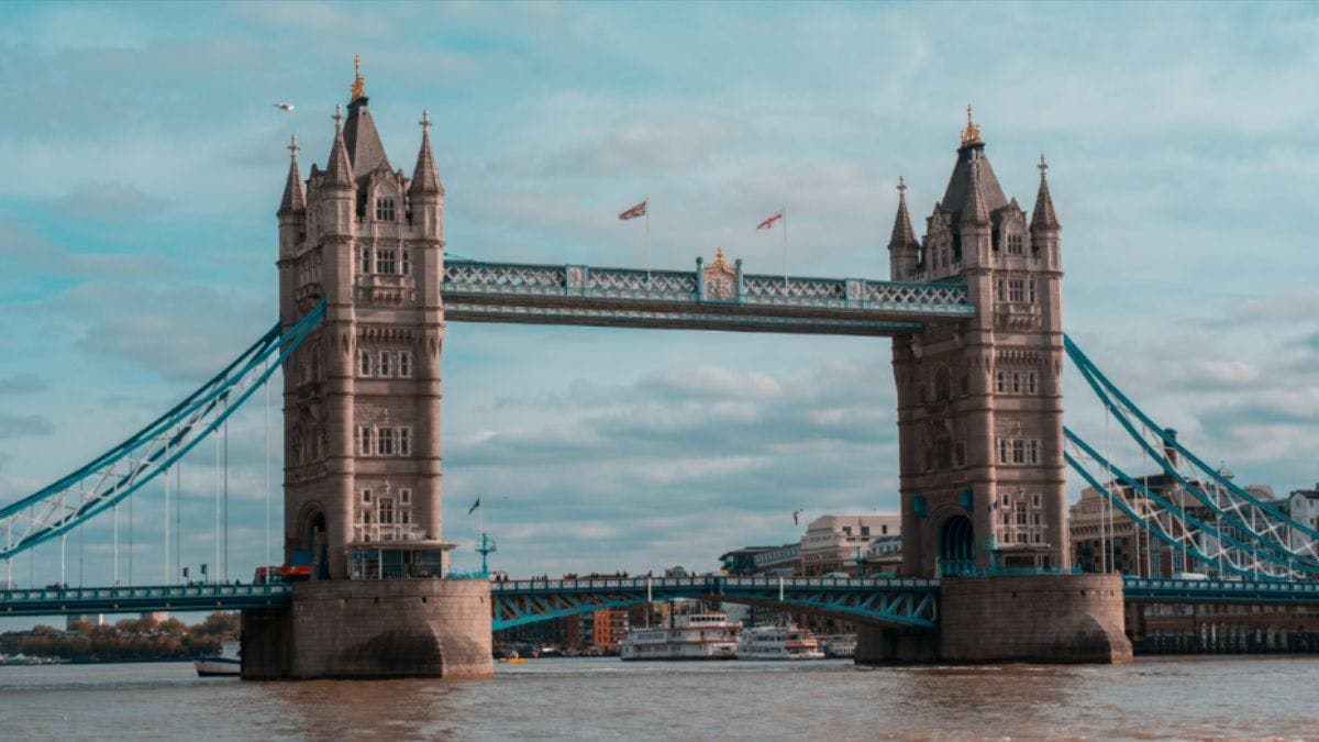 The Tower Bridge in London spans the River Thames, with a clear sky and flags flying atop the towers.